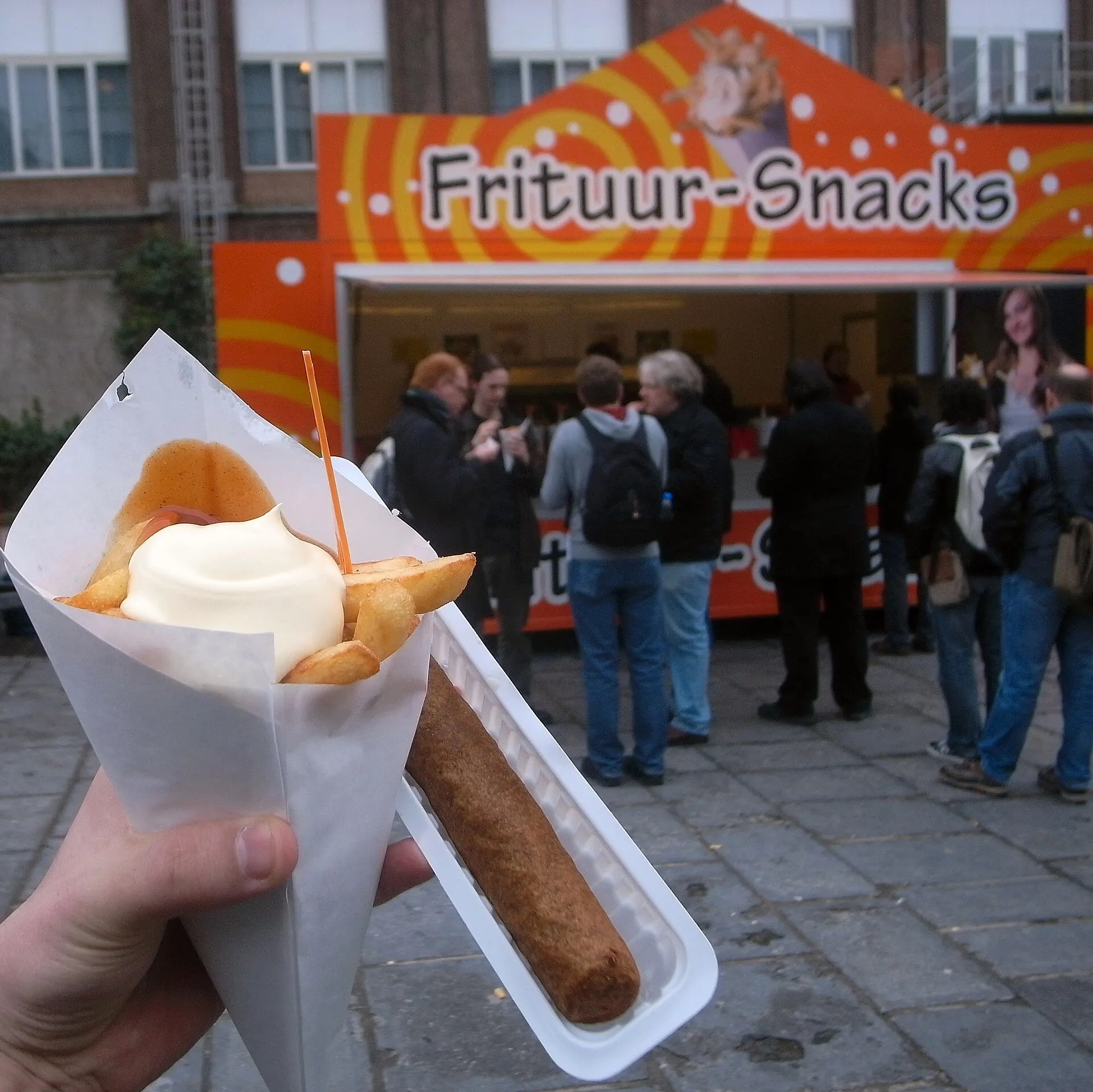 Un cornet de frites mayonnaise et une fricadelle devant une friterie